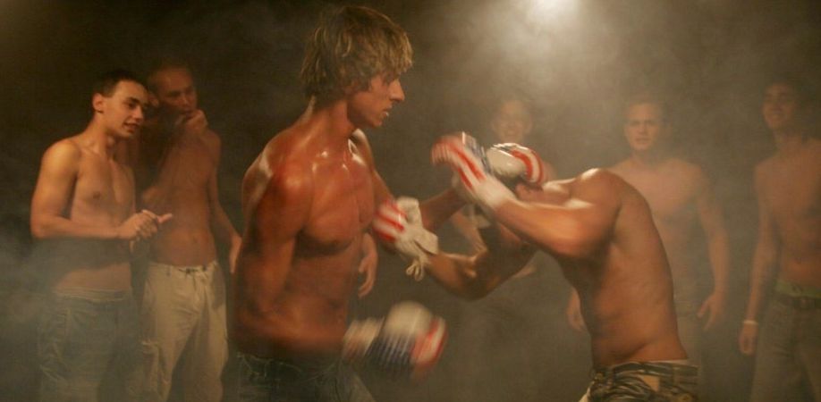 Two guys boxing in the dark with a crowd of shirtless guys watching them