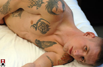 Shirtless, inked jock on bed, looking at camera.