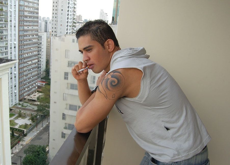 Inked young Latino takes a smoke break on the balcony